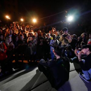 Facing the backs of two people lit up onstage, looking out at a crowd watching them in the dark