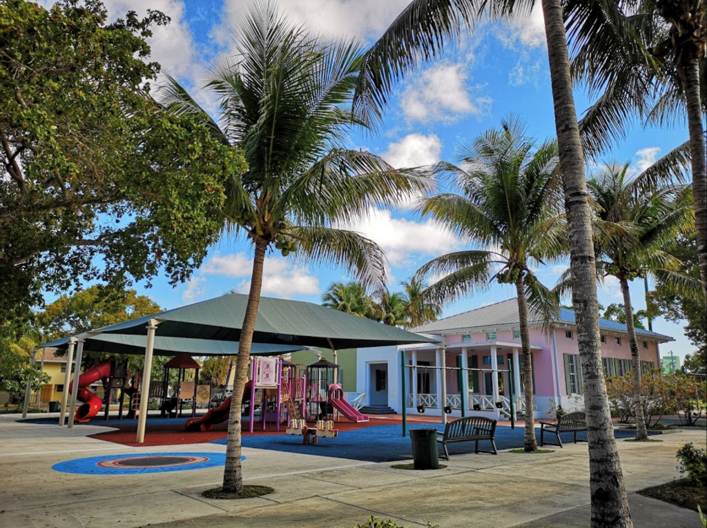A playground shaded overhead by a roof structure. It is surrounded by palm trees. There is a pink building in the background.