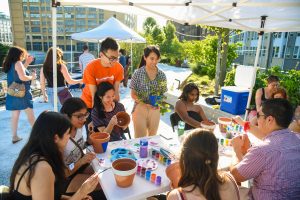 A group of people gathers around a table filled with art supplies, laughing and talking while painting flower pots.