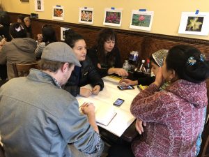 A crowded group of people sits and talks at a table inside a restaurant with pieces of paper in front of them. On the walls, there are taped printouts showing different flowers.