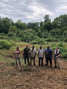 A group of 6 adults stand in front of a lot of grass and brush.