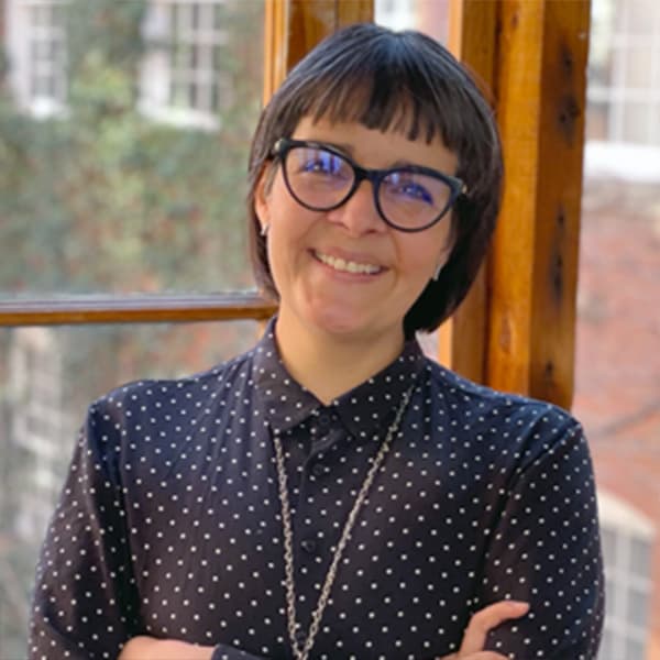 A woman with black hair and glasses, smiling warmly in a sun-lit window.