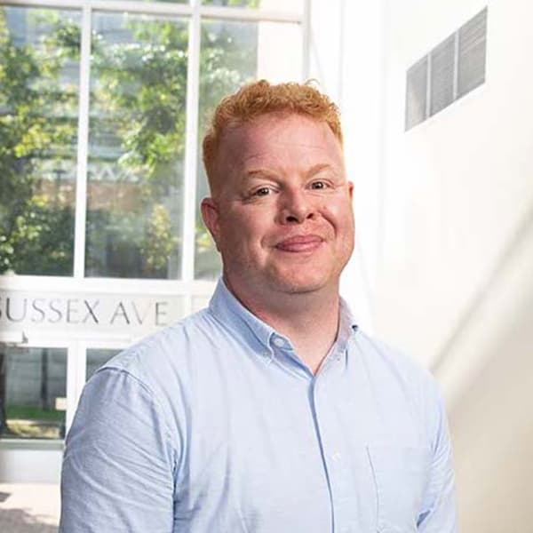 A blonde haired man in a blue oxford shirt smiling in a naturally lit lobby.