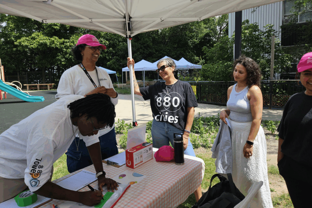 Group gathered under a tent at an outdoor table, with one person signing in on a clipboard while others stand nearby talking.