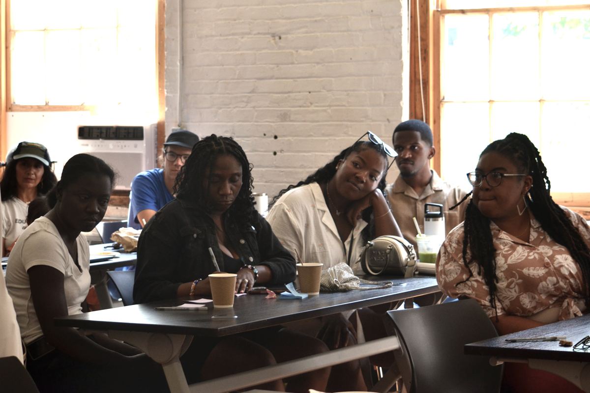 Participants sitting in a classroom-style setting with notebooks and drinks on the tables. The room has brick walls and large windows letting in natural light.