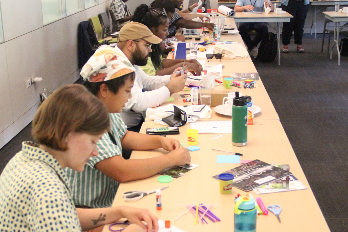 Participants seated at a long table covered with craft supplies, magazines, markers, and glue. They are engaged in a hands-on workshop activity or creative exercise.