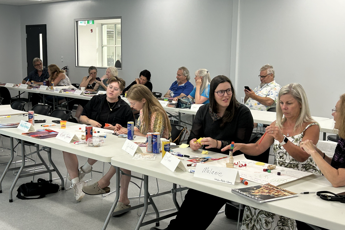 A group of people sit at long tables in a bright classroom participating in an arts-and-crafts workshop. The tables are covered with markers, glue sticks, paper, and other craft supplies. Name cards and drinks are placed in front of participants.
