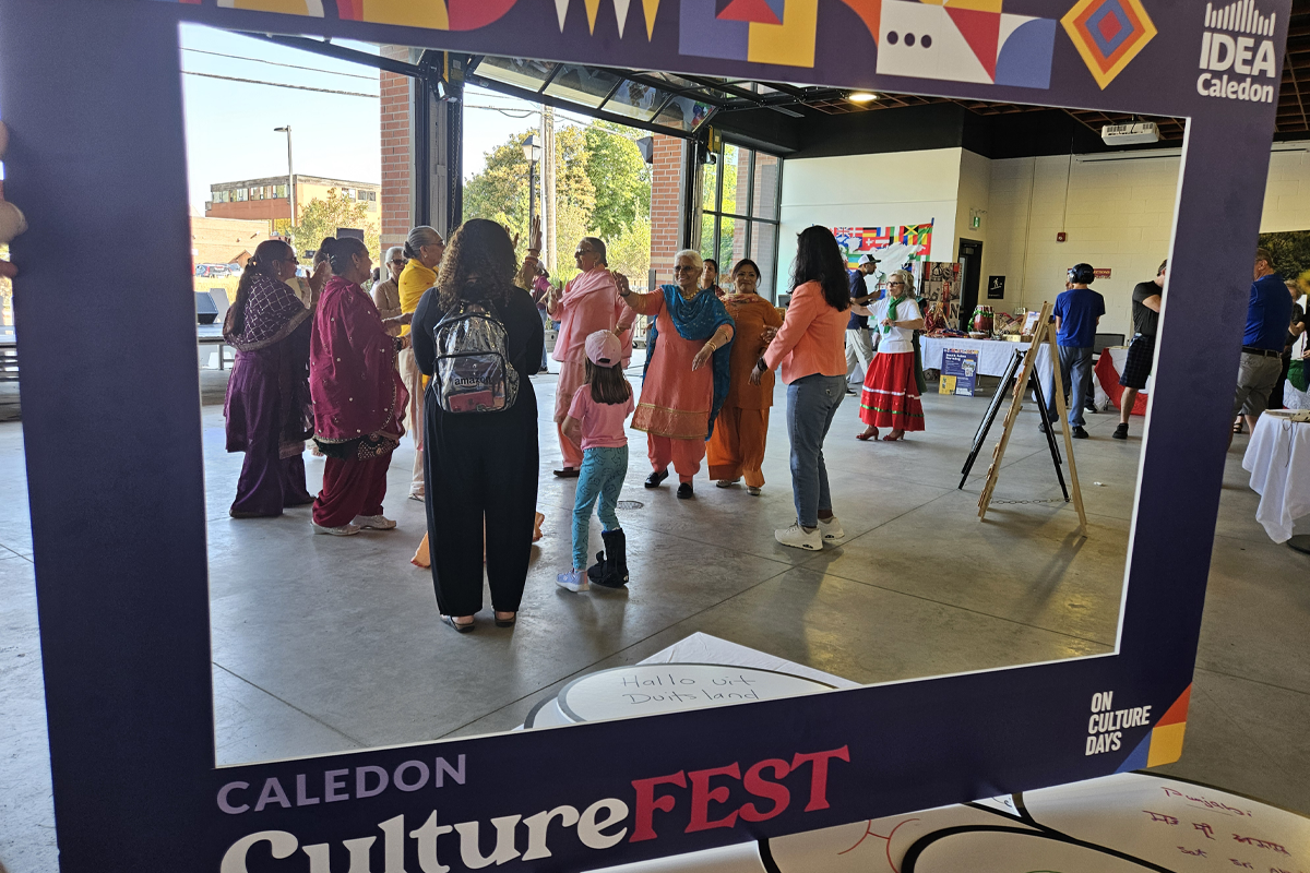 Large decorative frame labeled ‘Caledon CultureFEST’ stands in the foreground. Through the frame, an indoor cultural event is visible with people gathered in a circle participating in a dance activity. Several participants wear brightly colored traditional clothing. Display tables with artwork and information are arranged around the space. Large open doors at the back of the venue show trees and buildings outside.