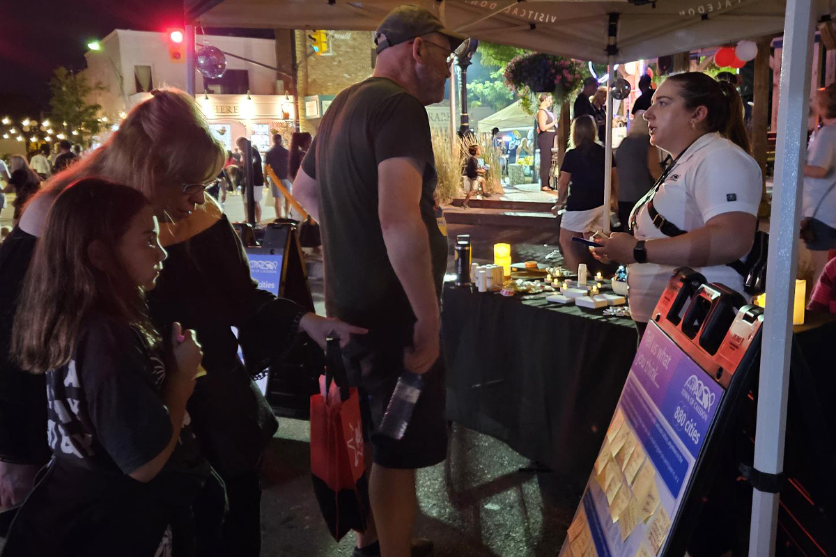 A crowd gathers at an outdoor nighttime street festival under string lights. Several people stand at a booth displaying small items on a table, while event signage and vendor tents line the street.