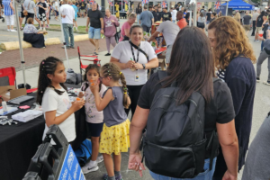 Children and adults gather at a busy outdoor event booth. The booth table is covered with supplies, while crowds fill the surrounding festival area with tents, signage, and activities.