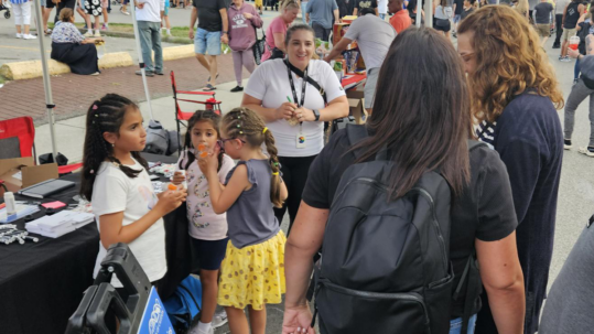 Children and adults gather at a busy outdoor event booth. The booth table is covered with supplies, while crowds fill the surrounding festival area with tents, signage, and activities.