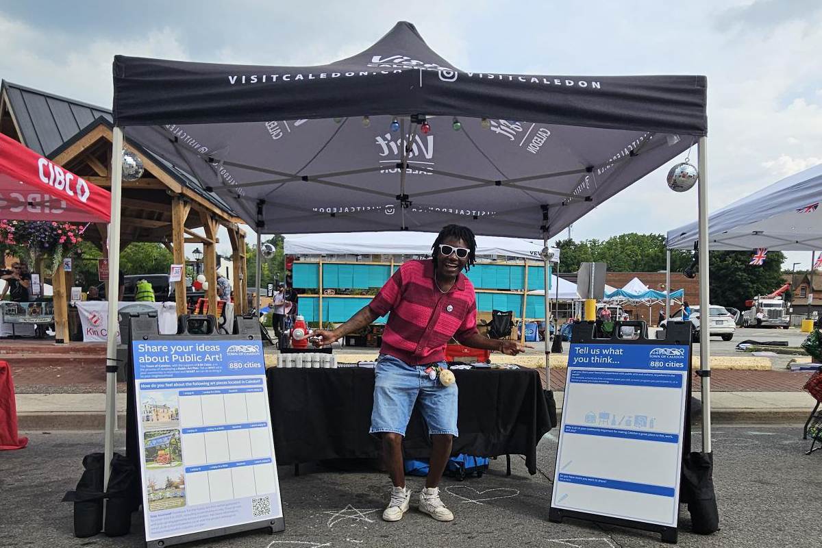 A person stands under a branded event tent at an outdoor festival. Two large display boards flank the tent, both inviting visitors to share ideas about public art. Vendor tents and festival-goers are visible in the background.