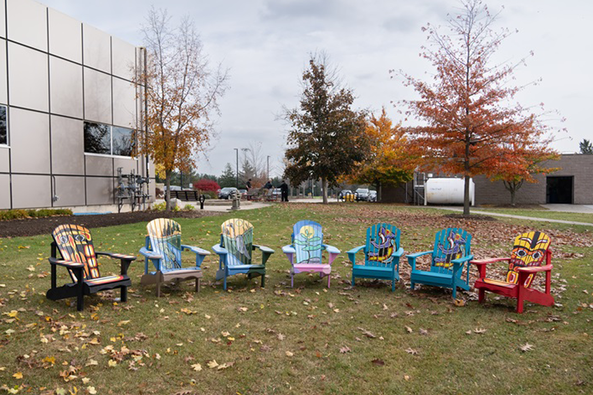 A row of seven brightly painted Adirondack chairs is arranged on a grassy lawn covered with scattered autumn leaves. Each chair features a different colorful artistic design. Behind the chairs, trees with red, orange, and yellow fall foliage stand near modern buildings with reflective windows.