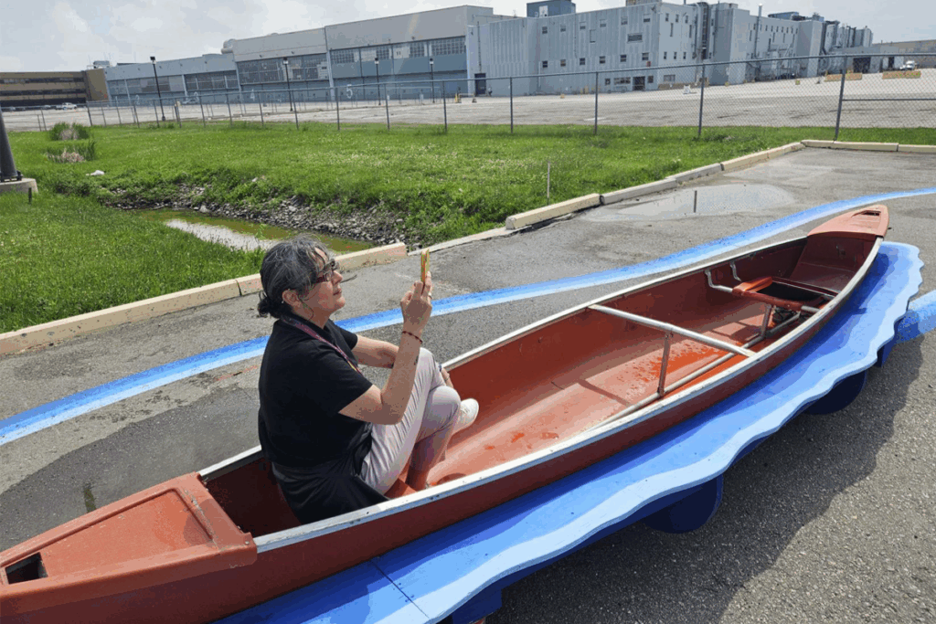 Person sitting in a canoe placed along a painted blue roadway, holding a pencil in an outdoor urban area.