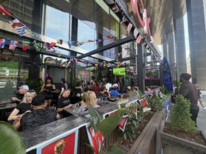 Patrons sit and stand on a busy downtown Toronto restaurant patio decorated with strings of international flags and FIFA World Cup-themed banners. A television screen broadcasts a soccer match as people gather outdoors between tall office towers.