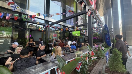 Patrons sit and stand on a busy downtown Toronto restaurant patio decorated with strings of international flags and FIFA World Cup-themed banners. A television screen broadcasts a soccer match as people gather outdoors between tall office towers.