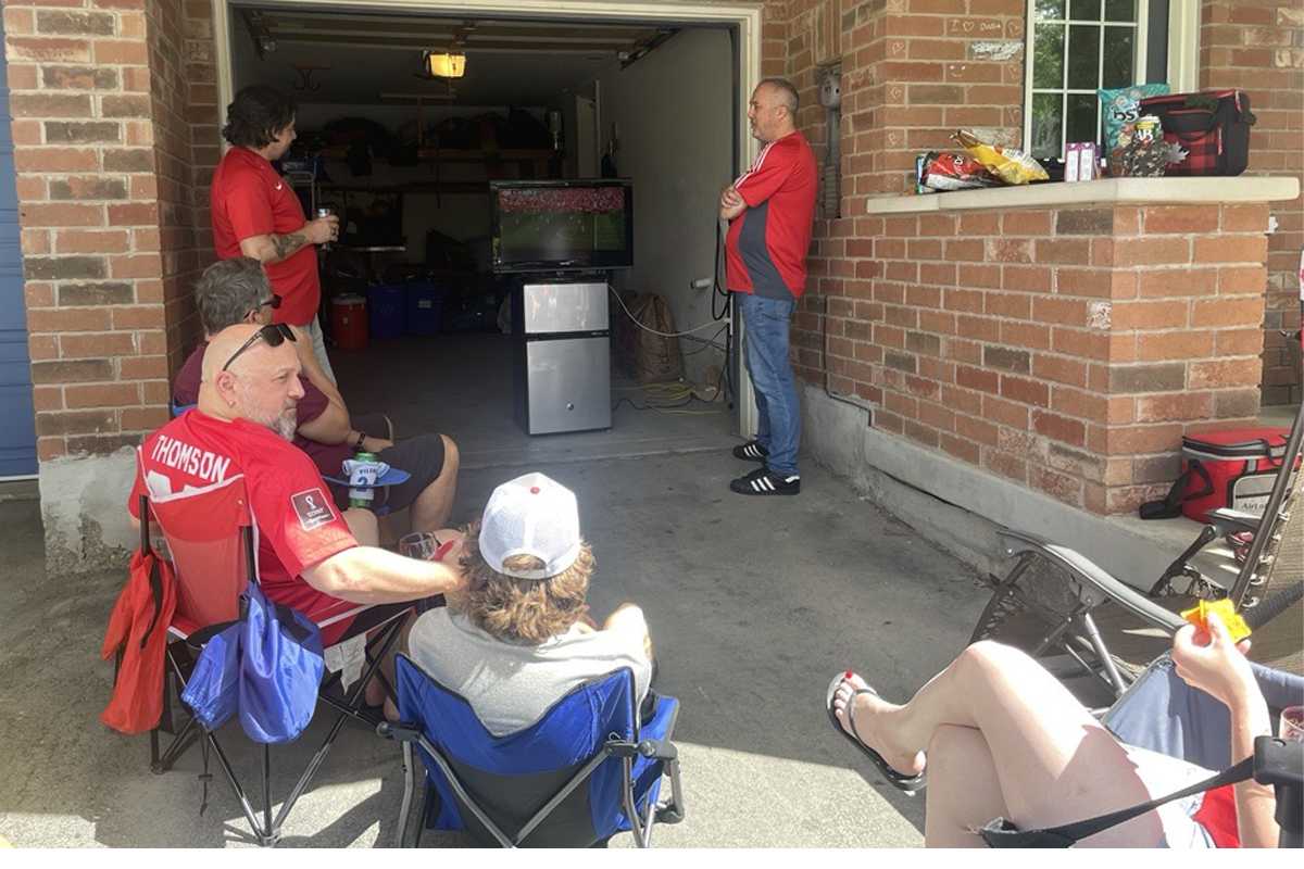Neighbours gather in a residential driveway to watch a soccer match on a television set up in an open garage. People sit in folding chairs and stand nearby, creating a shared outdoor viewing experience in the neighbourhood.