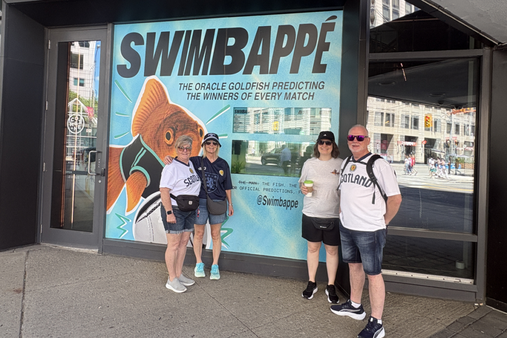 Four people wearing Scotland football jerseys pose in front of a large “SWIMBAPPÉ” storefront display featuring a goldfish mascot and the slogan “The Oracle Goldfish Predicting the Winners of Every Match.”