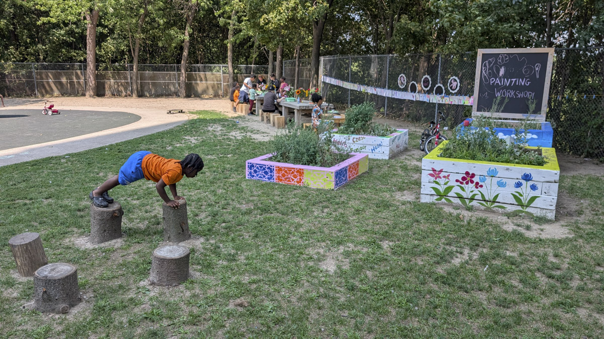A child in an orange shirt planks across wooden logs on a grassy lawn, with painted garden beds and a "Painting Workshop" chalkboard sign visible in the park background.